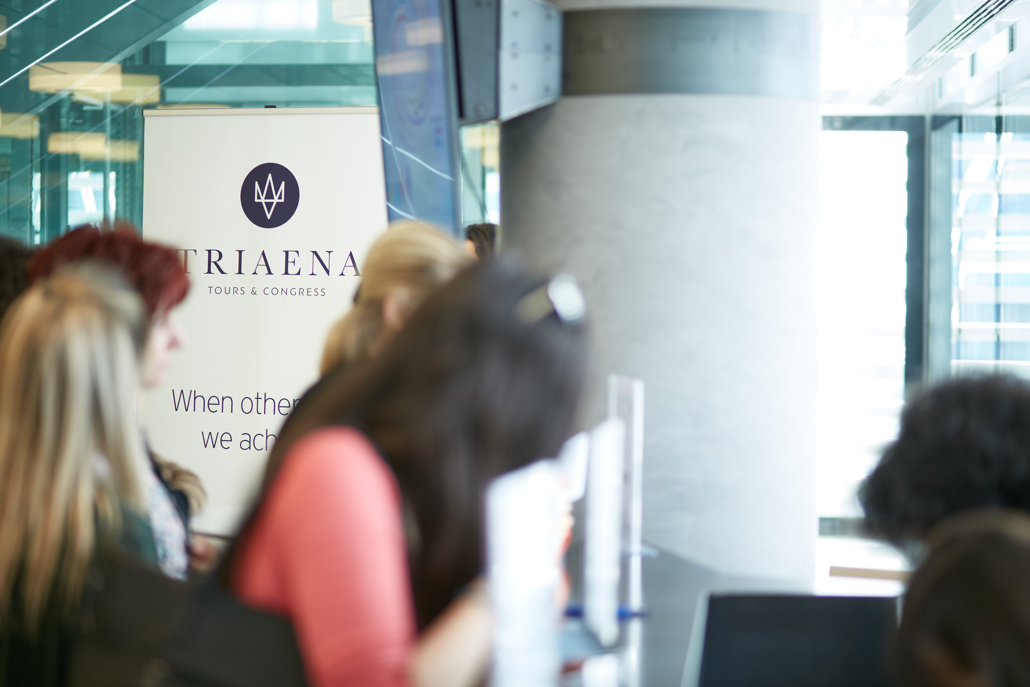 Onsite secretariat area during a busy day of conference, participants lined up to get their name tags – badges in a big modern venue, employs working, and a roll up banner on the back showing Triaena with their logo and moto When others try, we achieve! 